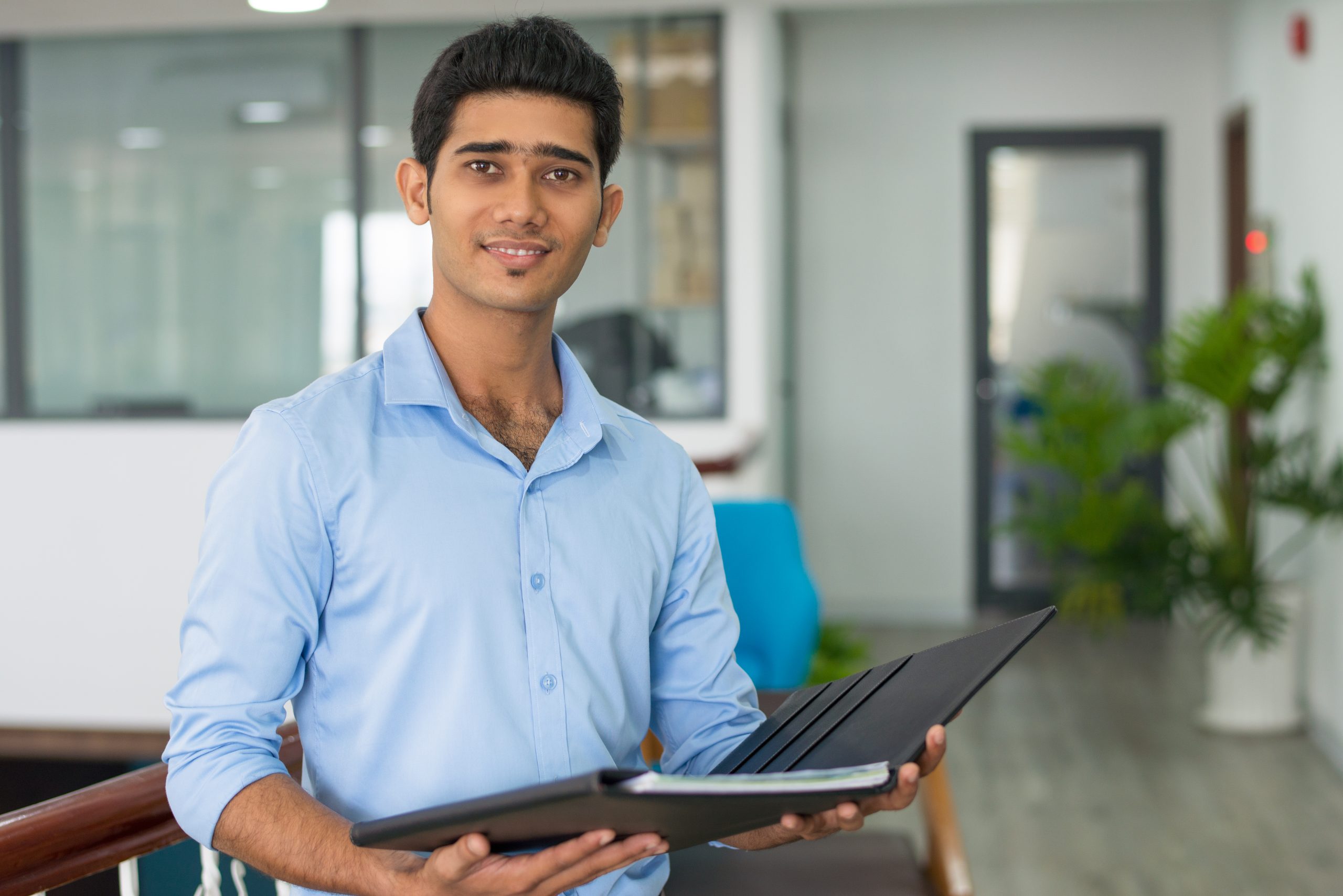 Positive Indian financial advisor holding open folder with documents and looking at camera. Smiling confident successful young businessman working in office. Business analyst concept